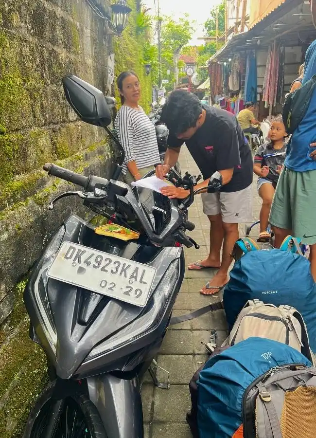 Viajeros en scooter gris firmando papeles en calle con mochilas.