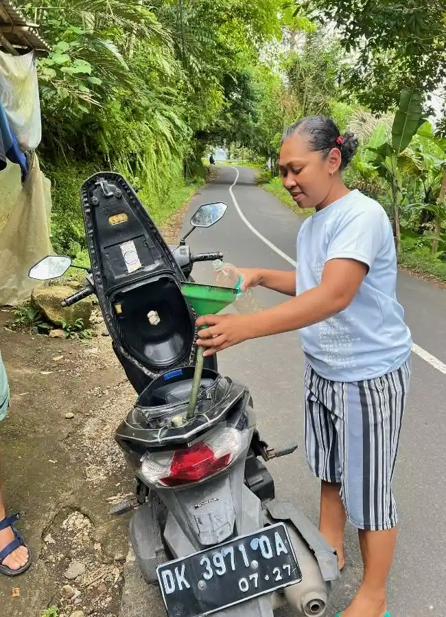 Mujer repostando scooter con botella y embudo en entorno rural.