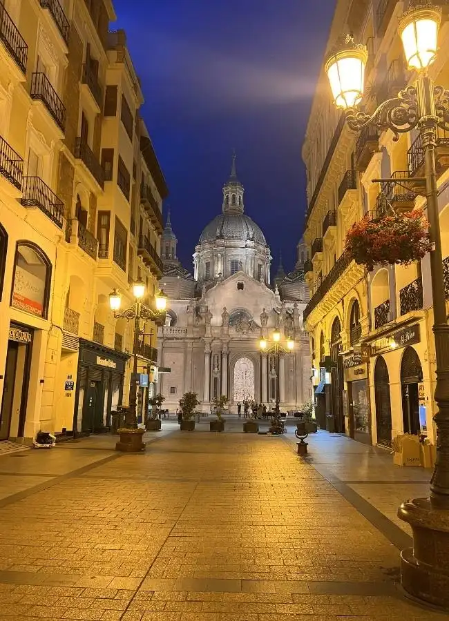 Basílica del Pilar iluminada al anochecer desde calle adoquinada.