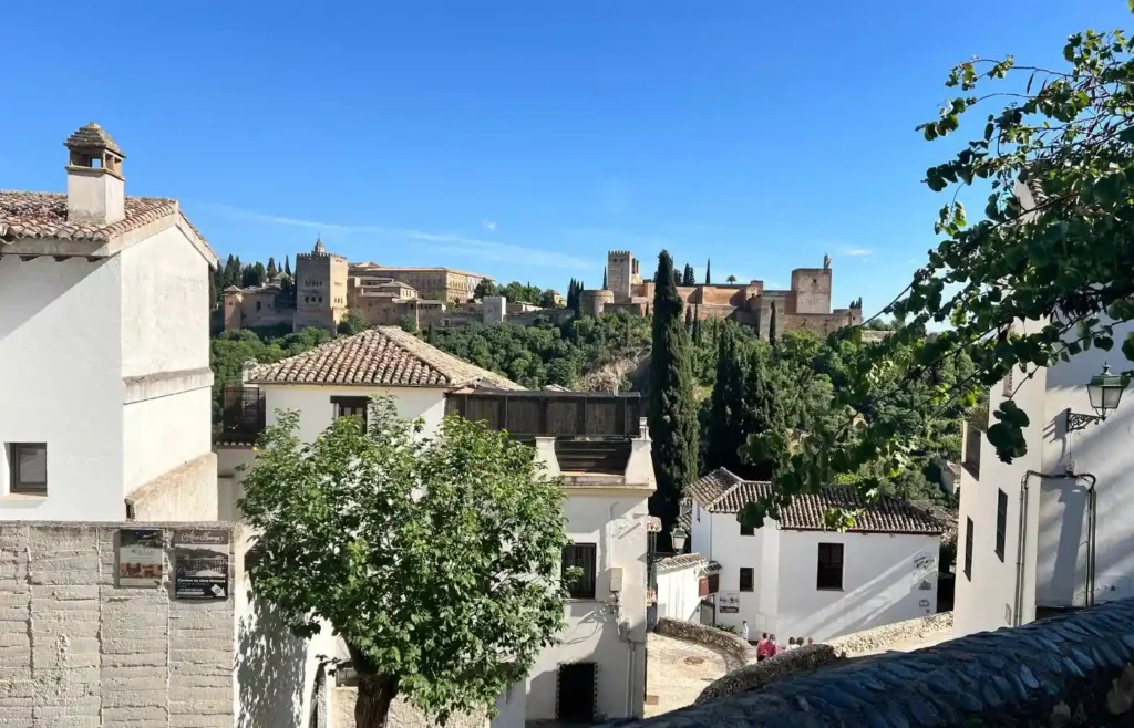 Casas blancas y calle empedrada del Albaicín con la Alhambra sobresaliendo entre la vegetación.