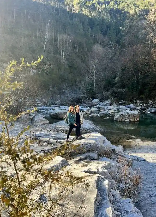 Dos mujeres en rocas heladas junto a un río boscoso.