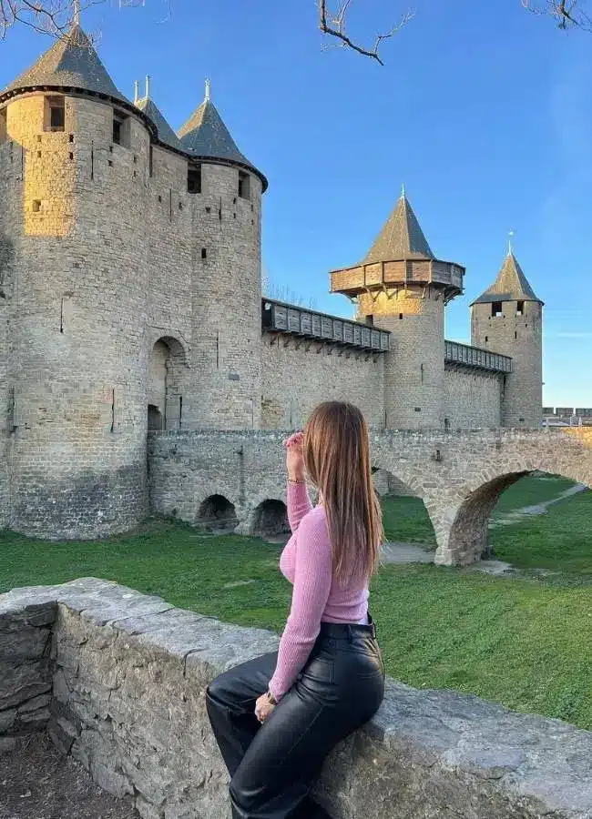 Mujer admirando castillo medieval amurallado bajo cielo azul.