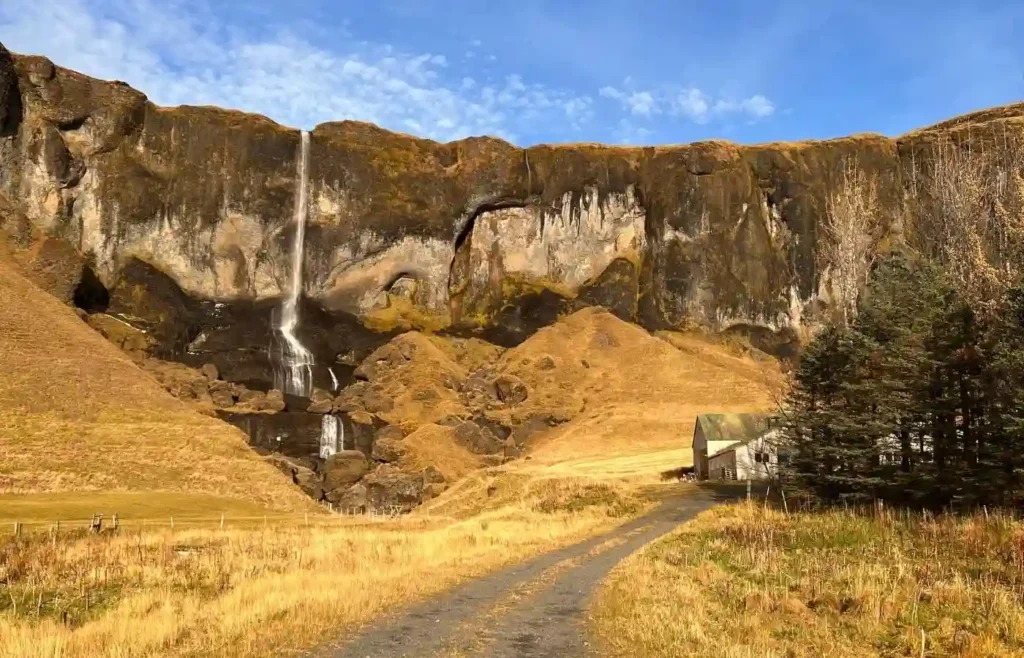 Cascada alta, acantilado, campos dorados y casa solitaria.