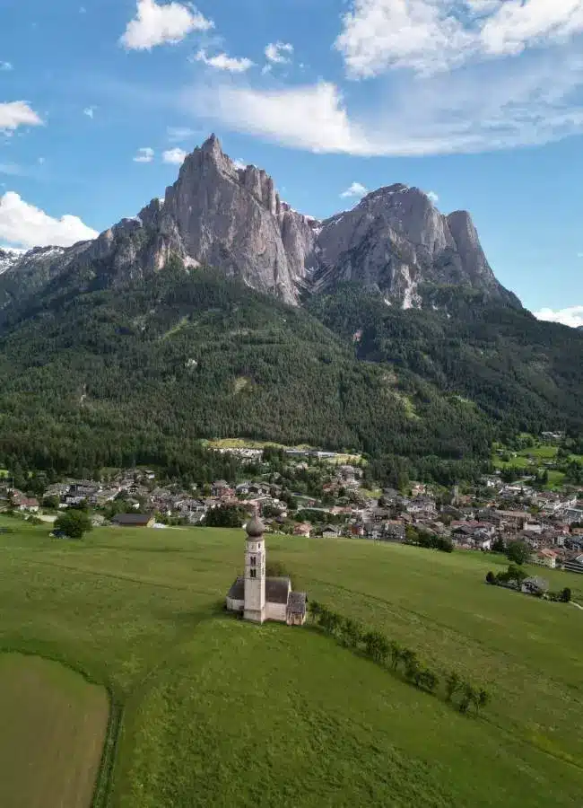 Iglesia, pradera, pueblo y montañas majestuosas bajo cielo azul.