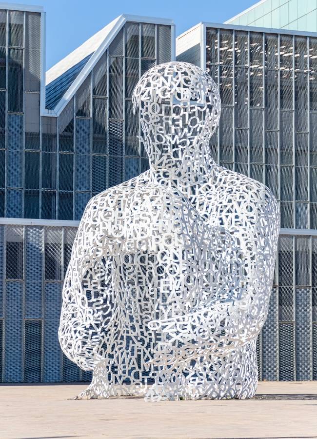Escultura hombre de letras Jaume Plensa con edificios modernos.