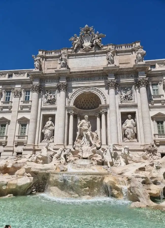 Vista frontal de las esculturas de mármol y las aguas de la Fontana di Trevi bajo un cielo azul intenso.