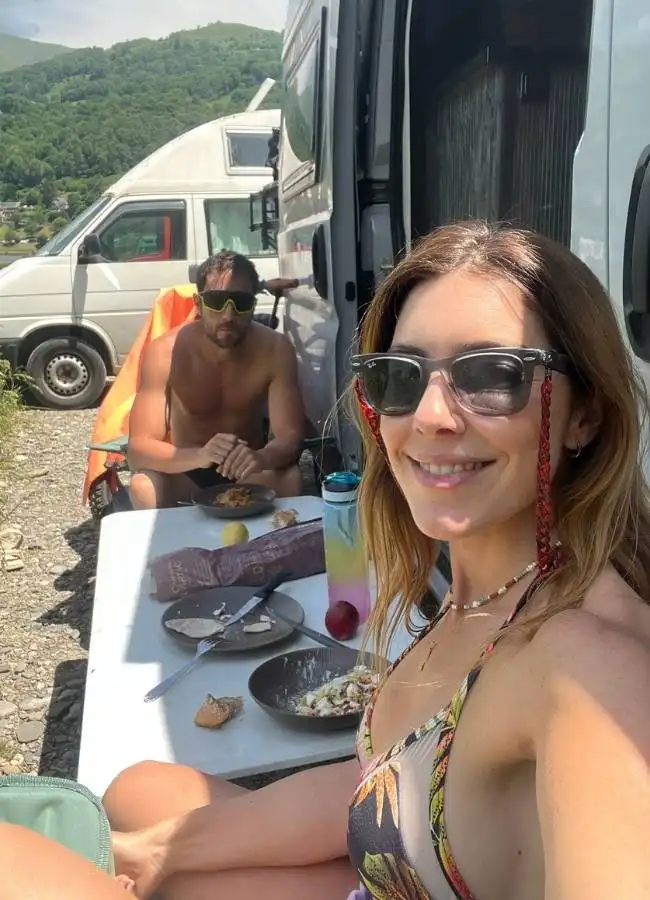 Pareja comiendo al aire libre con furgonetas camper y montañas.