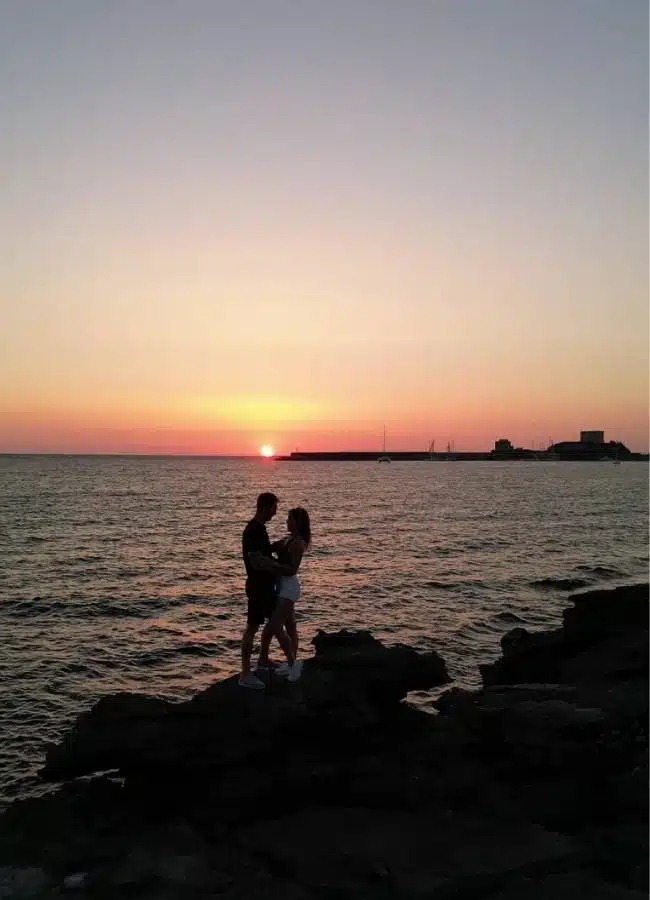 Pareja contemplando la puesta de sol sobre el mar desde unas rocas en Cerdeña.
