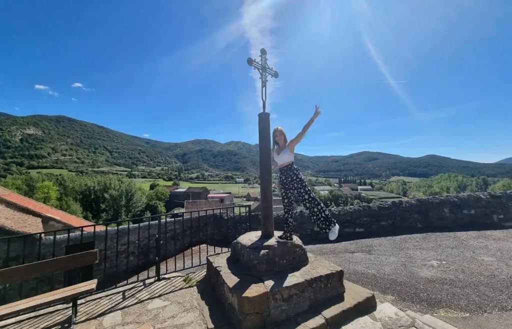 Mujer posando junto a una cruz de piedra en un mirador con vistas a las montañas de Echo