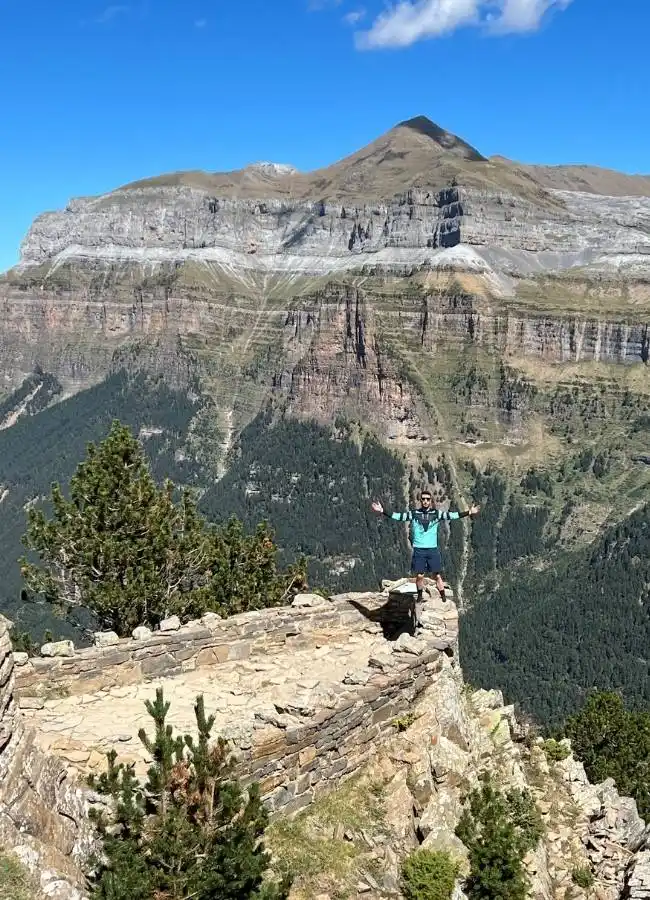 Hombre en cima de montaña con brazos extendidos, paisaje pirenaico.