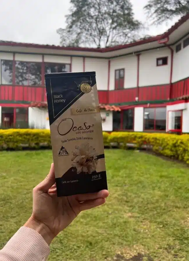 Mano sosteniendo un paquete de café de especialidad frente a la casona de la Finca El Ocaso.