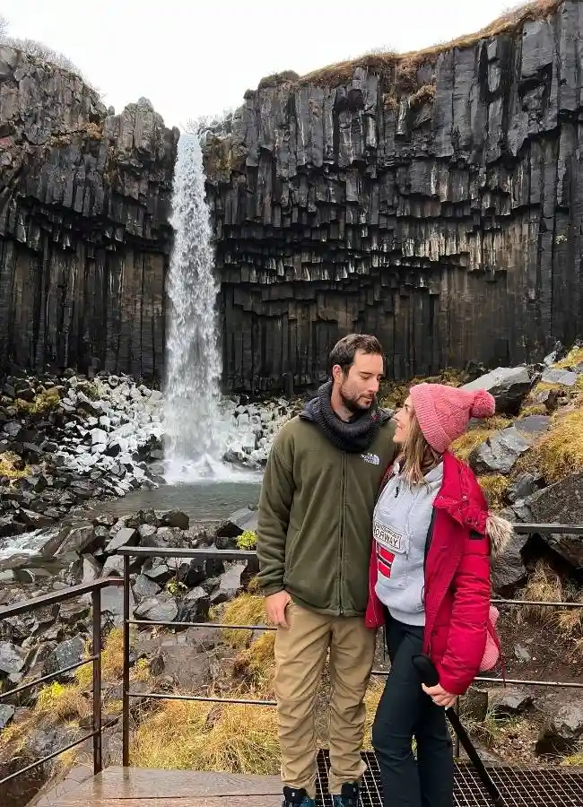 Pareja de viajeros posando juntos frente al anfiteatro de basalto de la cascada Svartifoss.