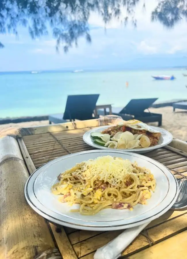 Espagueti carbonara y nasi goreng en mesa de bambú junto al mar.
