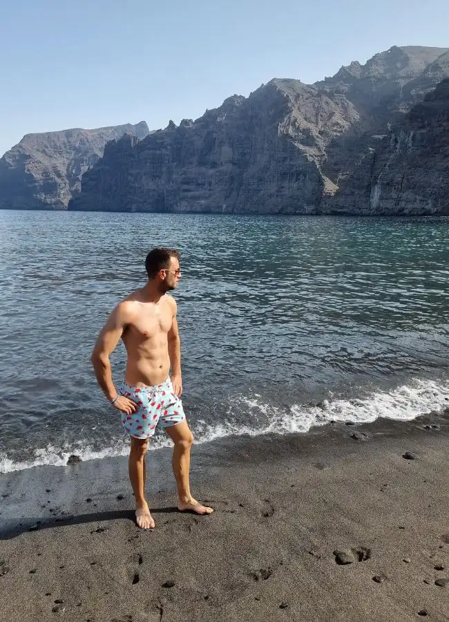 Hombre caminando por una playa de arena volcánica negra bajo los acantilados.