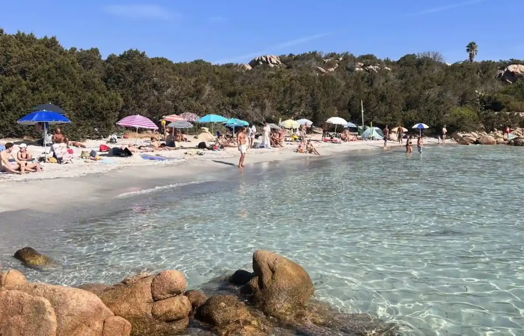 Playa con gente, sombrillas y mar transparente bajo cielo azul.