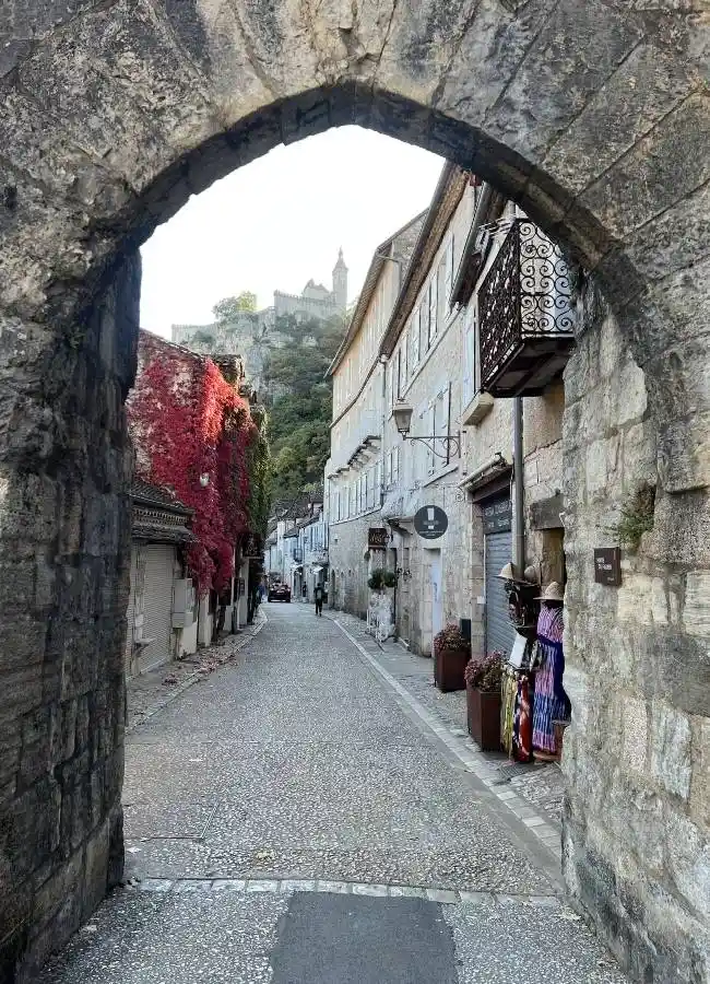 Calle adoquinada con arco de piedra, edificios antiguos y castillo.