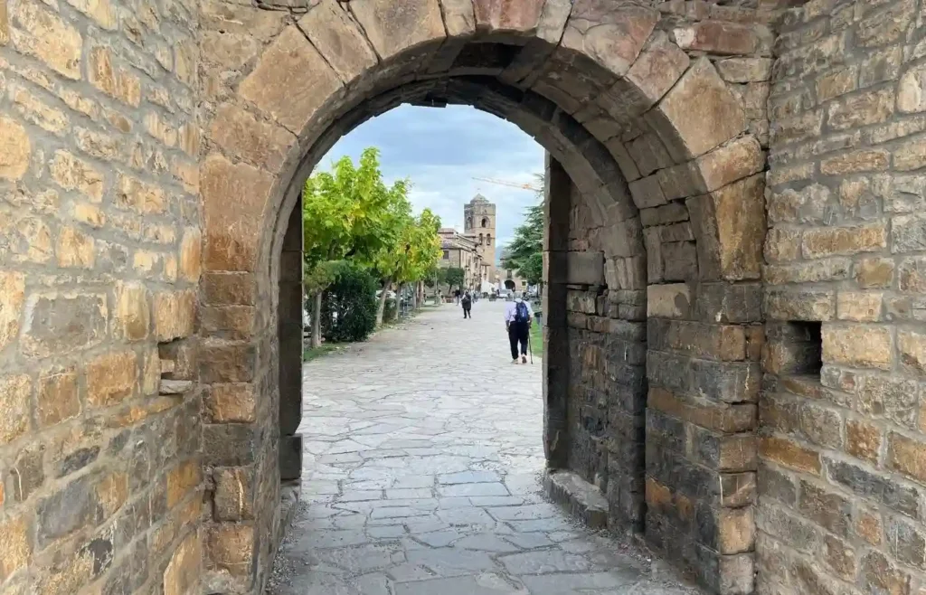 Entrada por un arco de piedra a la plaza mayor de Aínsa con turistas paseando.