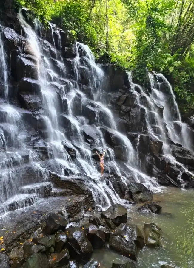 Mujer en bañador posa feliz ante impresionante cascada tropical.