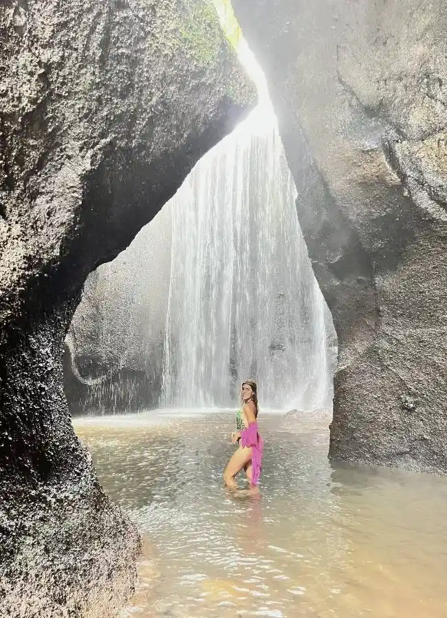 Mujer bañándose dentro de una cueva iluminada por el sol con una cascada al fondo en Bali.