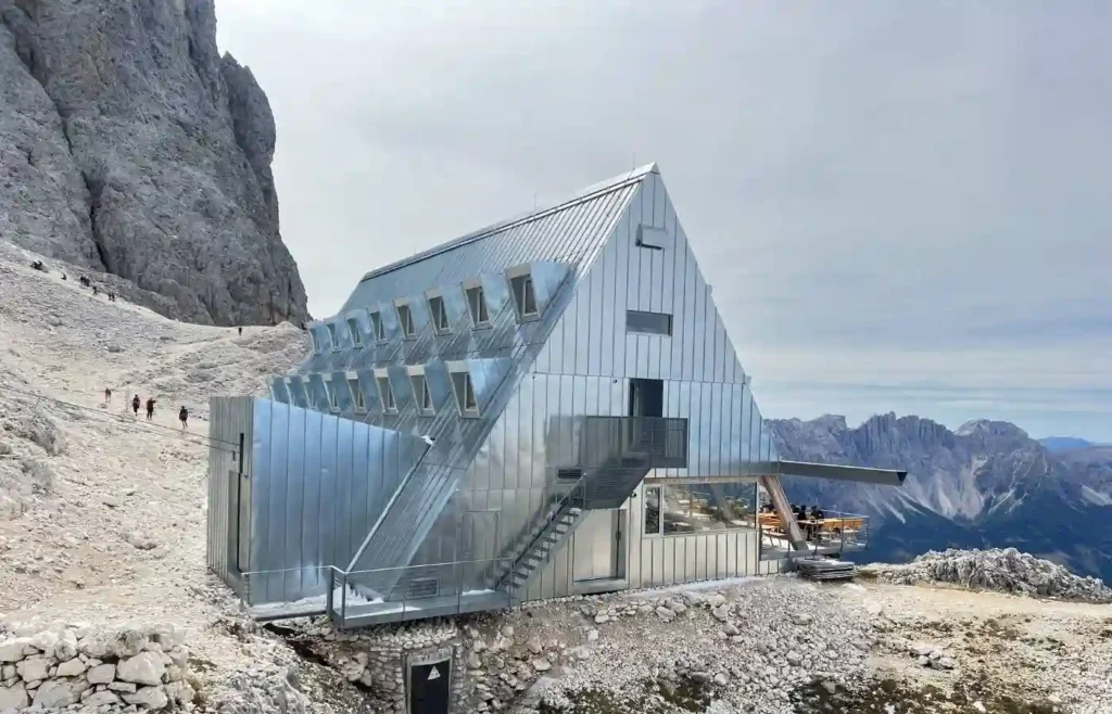 Moderno refugio alpino metálico en montaña rocosa con senderistas.