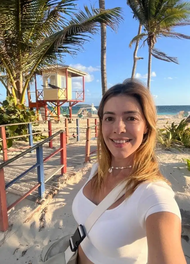 Selfie de mujer sonriendo en una playa soleada con palmeras al fondo.