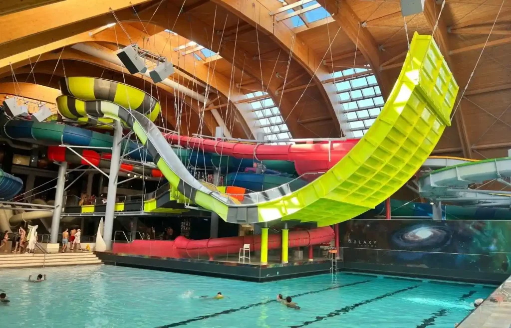 Parque acuático interior con toboganes coloridos y piscina llena de gente.