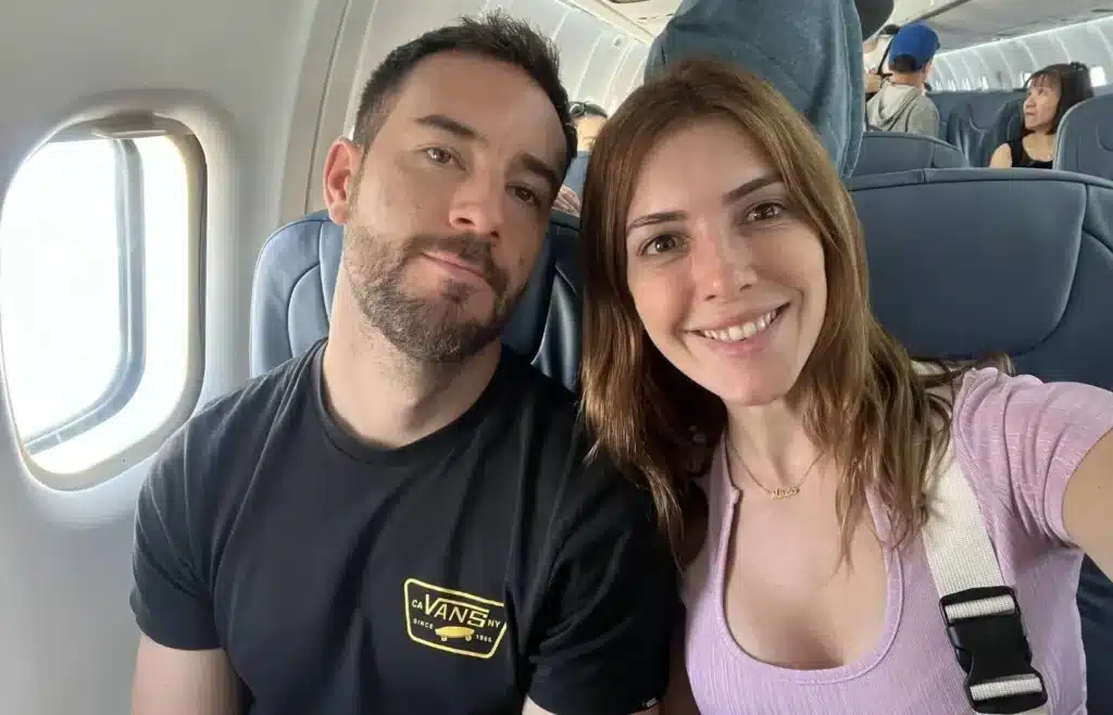 Héctor y Mireia sentados en sus asientos junto a la ventanilla del avión sonriendo al comenzar su viaje.