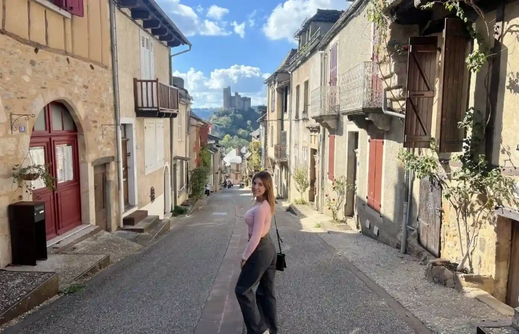 Mujer sonriente en calle adoquinada con castillo medieval al fondo.