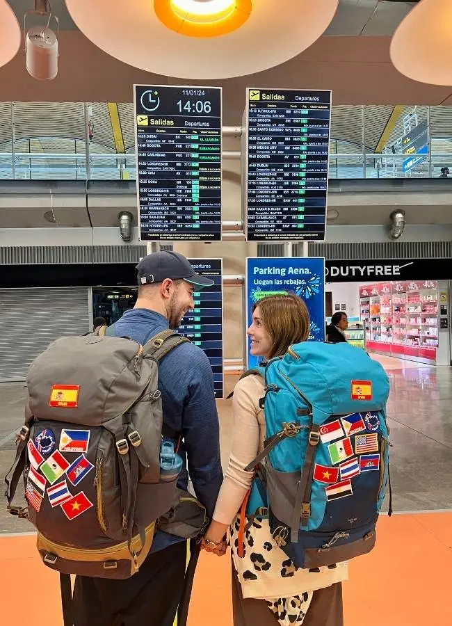 Mireia y Héctor de Viajeros con Ruta de espaldas mirando los paneles de salidas de vuelos en el aeropuerto con mochilas de viaje llenas de parches de países.