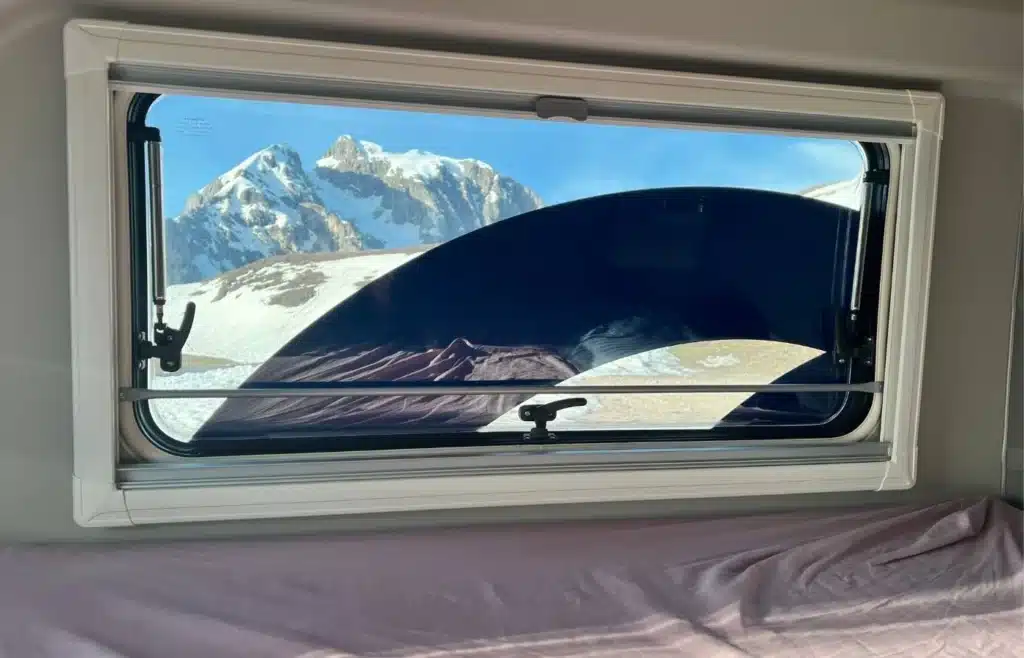 Ventana de caravana con vistas a montañas nevadas y persiana.